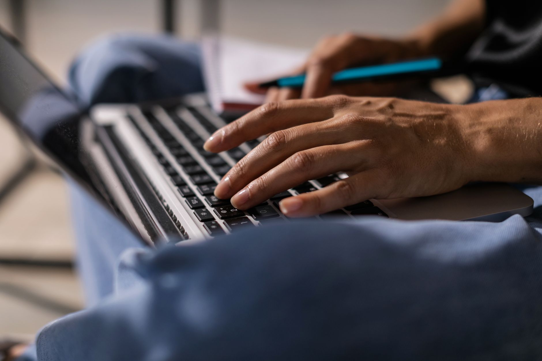A writer types on a laptop, a pen and notes in hand