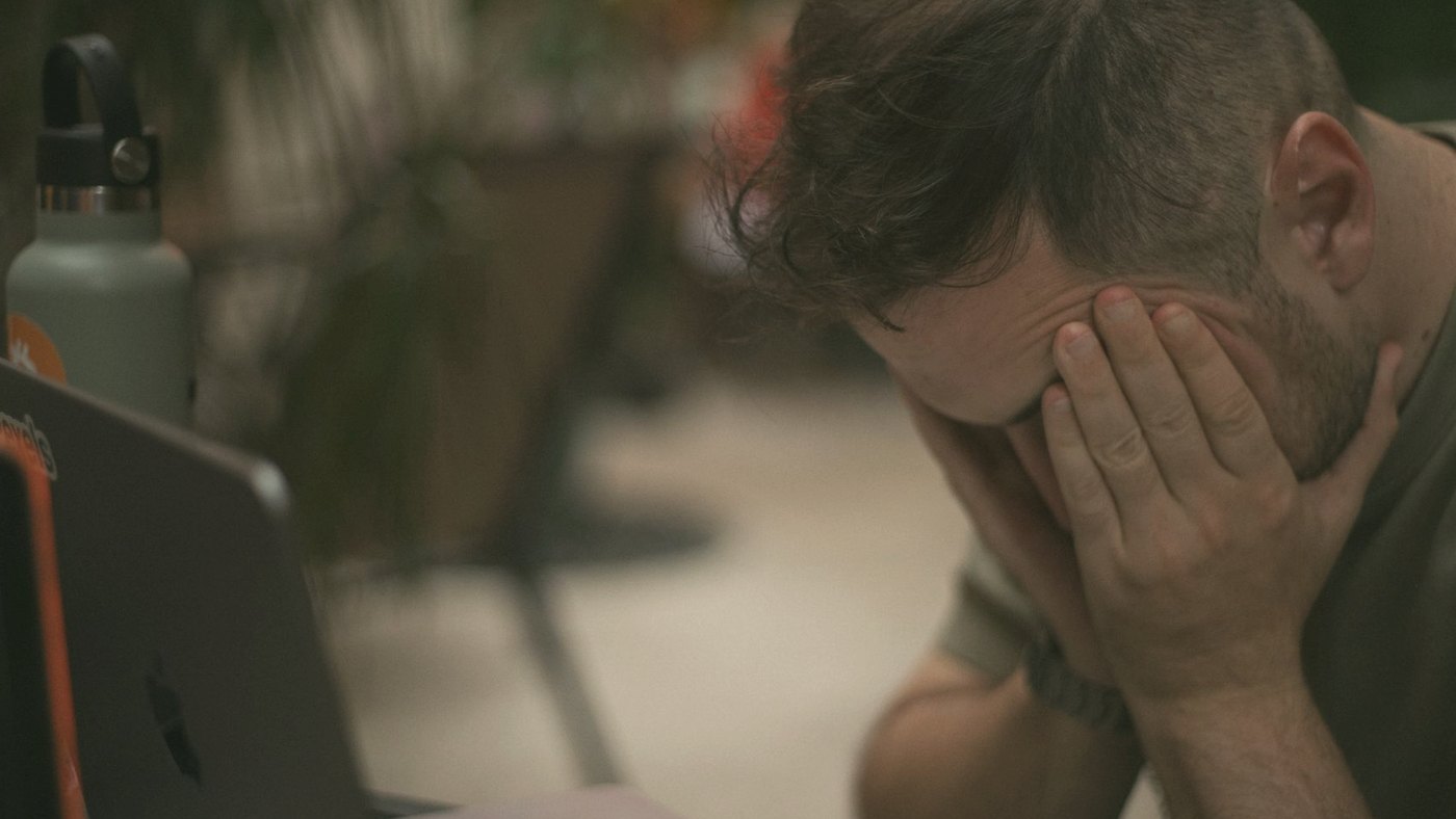 Man appears stressed, hands on his face beside a laptop